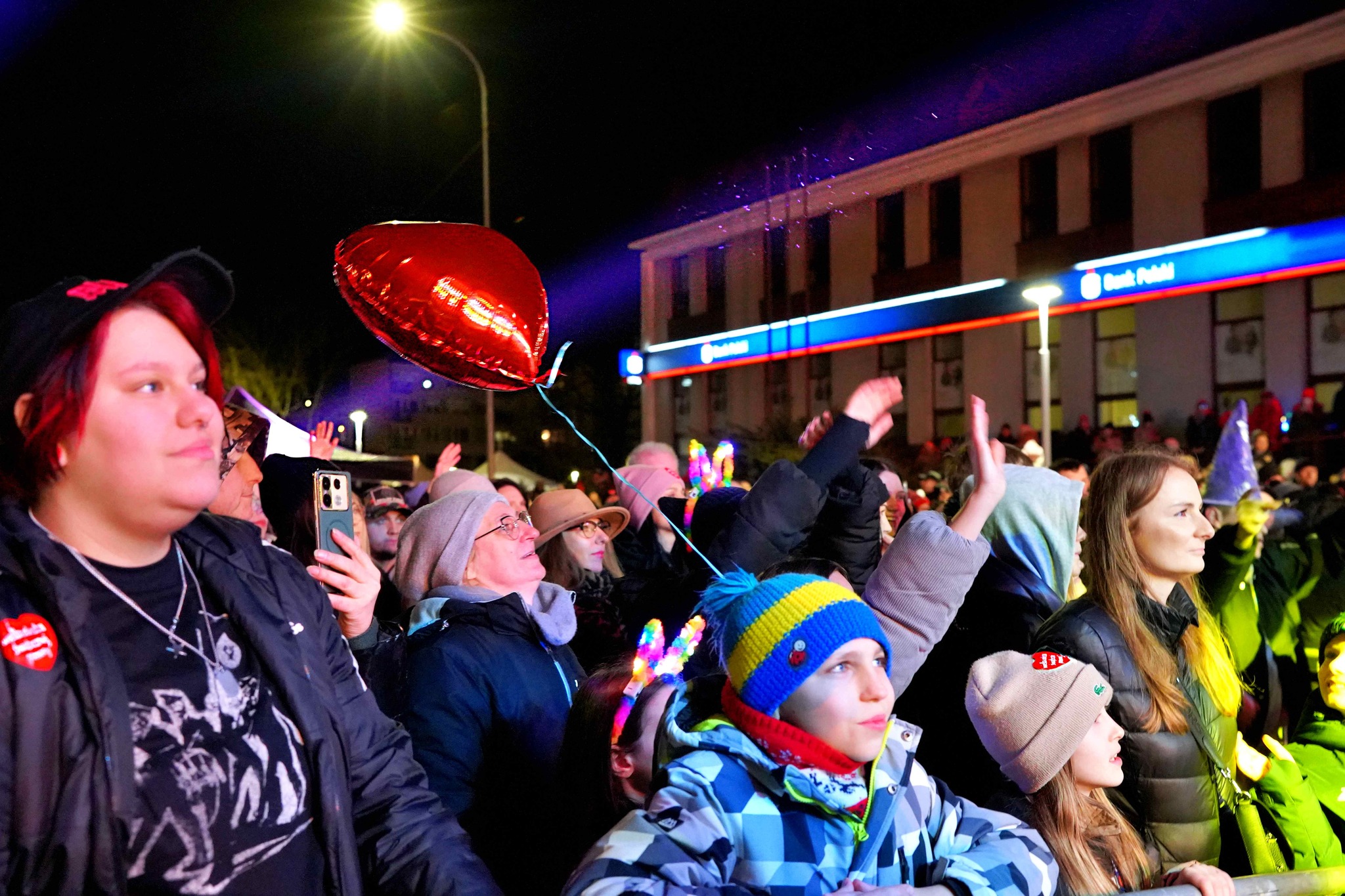 Zdjęcie przedstawia tłum ludzi podczas koncertu WOŚP w Kole. Uczestnicy są ubrani ciepło, widać kolorowe światła, balon w kształcie serca i entuzjastyczne reakcje publiczności. W tle widoczny jest budynek banku.