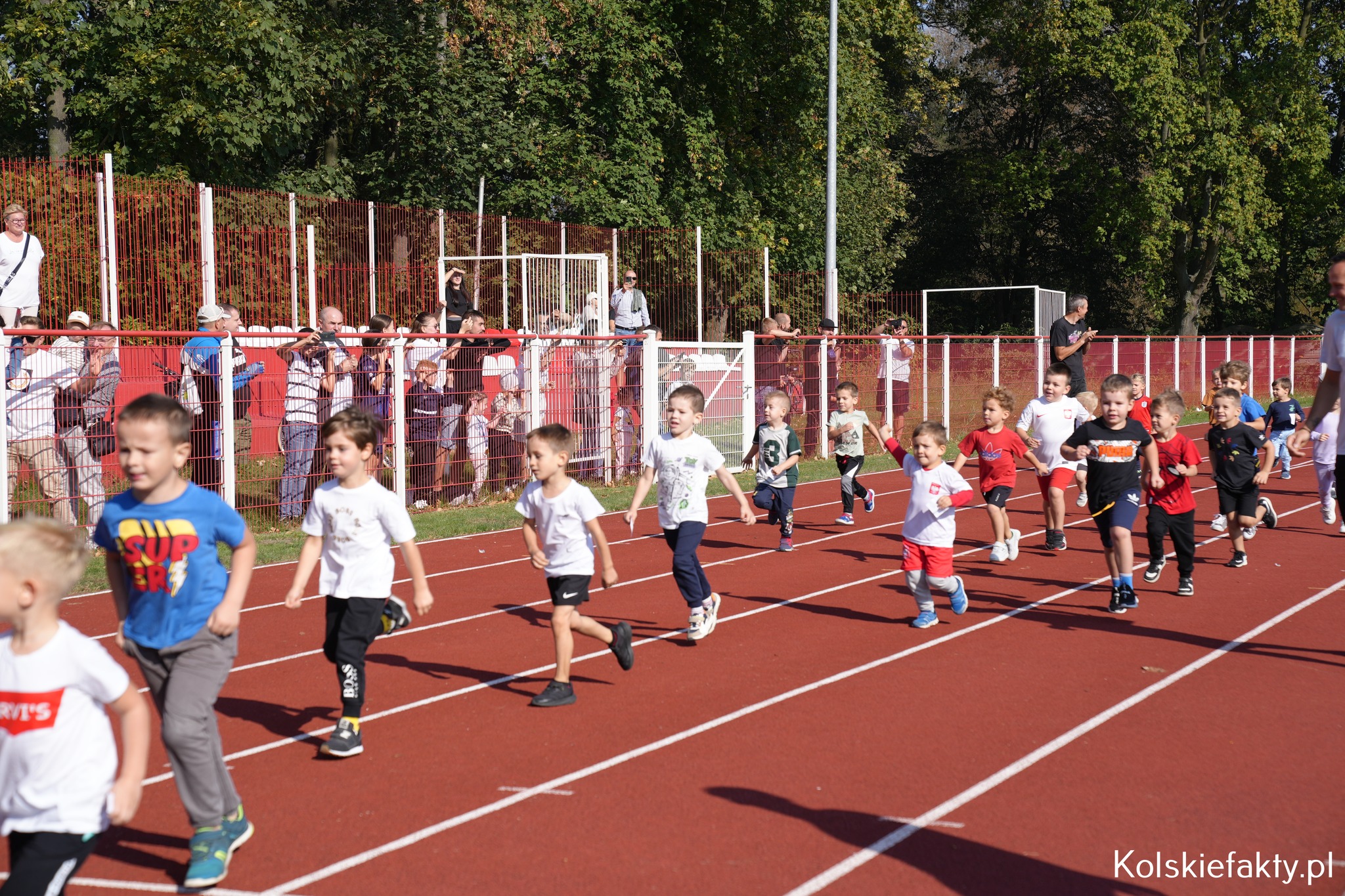 Zdjęcie przedstawia grupę małych dzieci biegnących na bieżni wokół stadionu. W tle widać rodziców przy ogrodzeniu. Pogoda jest słoneczna, a dzieci uczestniczą w zawodach sportowych w ramach 44. Międzynarodowego Biegu Warciańskiego 